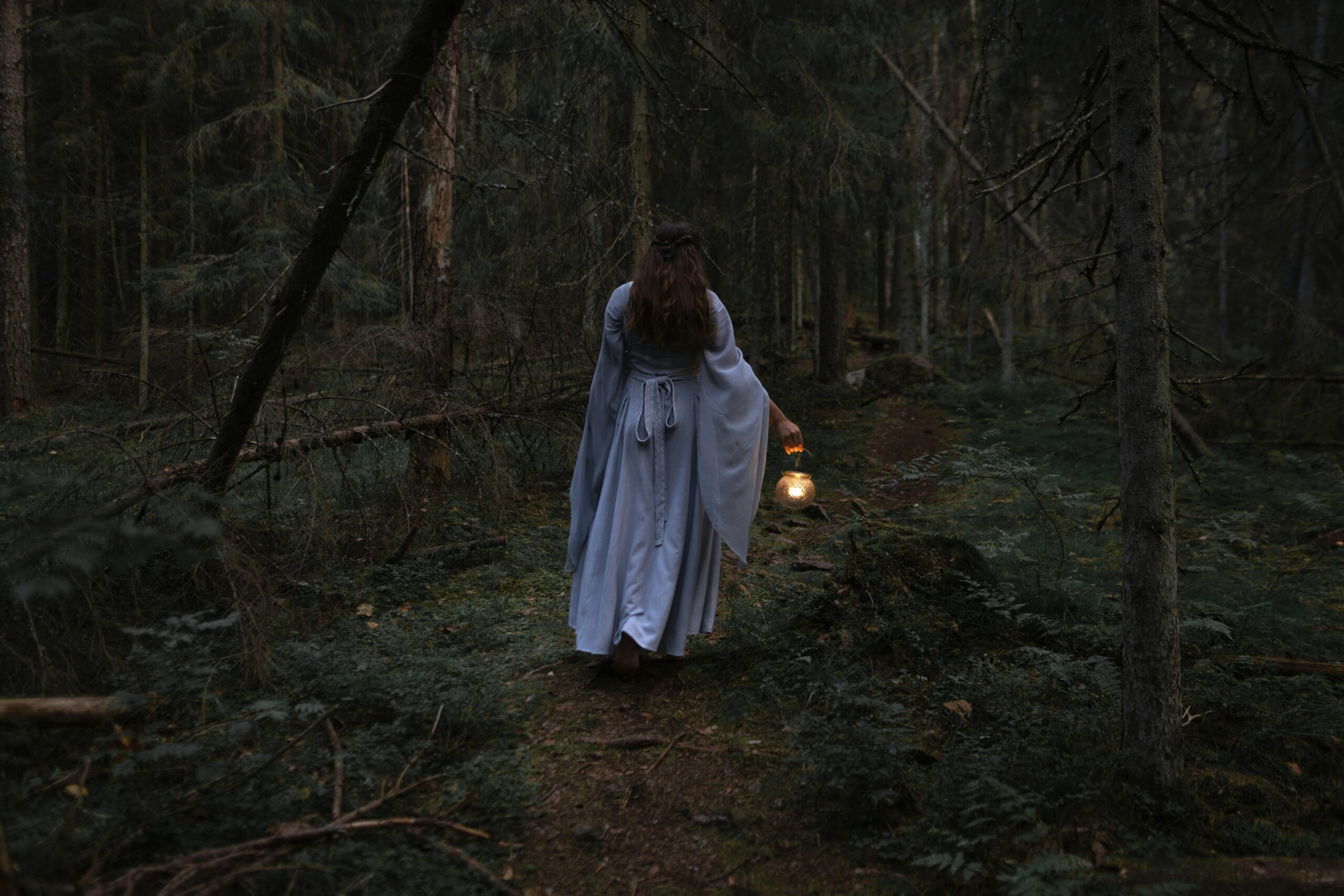 A person in a white cloak carrying a lantern through a dark forest