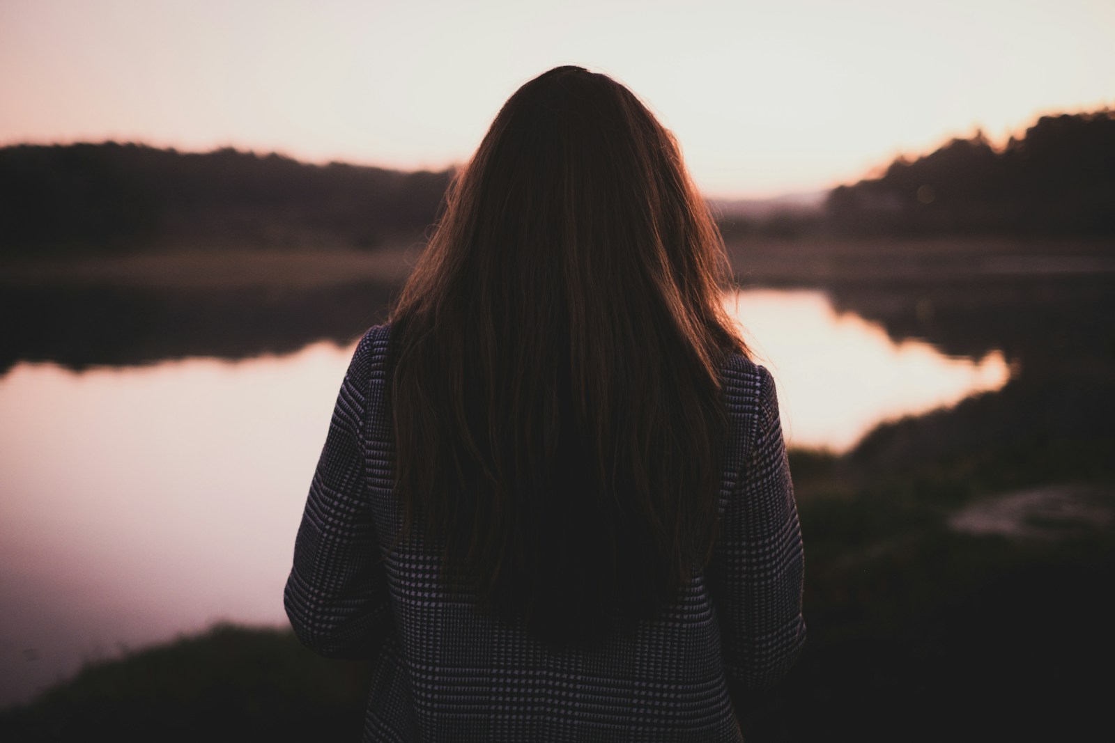 A woman with long hair, seen from behind in a checked shirt, looking out over a still lake at dusk.
