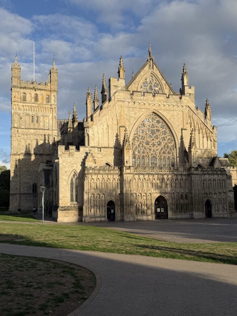 The west front of Exeter Cathedral lit by evening sun, with the green in front