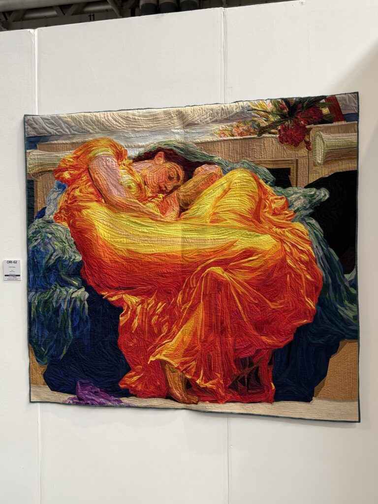 A quilt recreating the painting Flaming June, a sleeping woman in a glowing orange dress, hung on a white display wall
