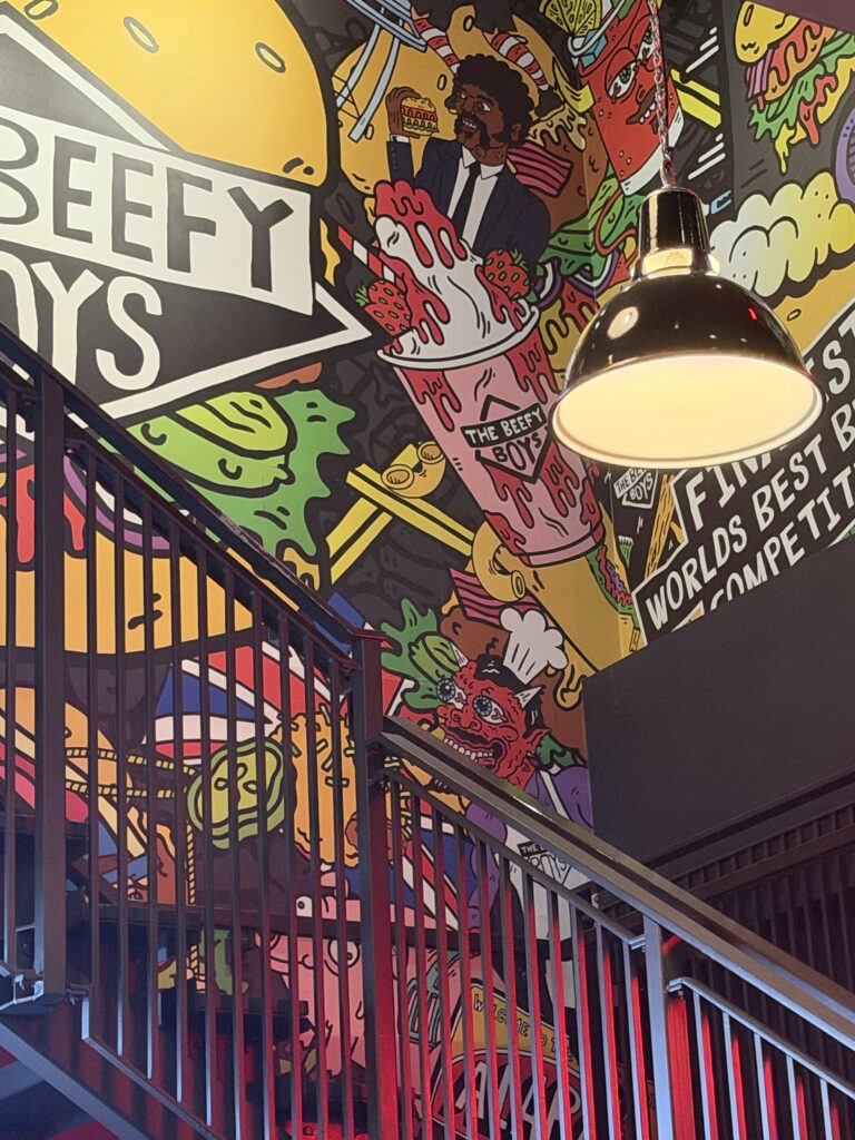 A colourful cartoon mural covering the stairwell wall at The Beefy Boys in Exeter, with the restaurant's logo, burgers and a dripping milkshake